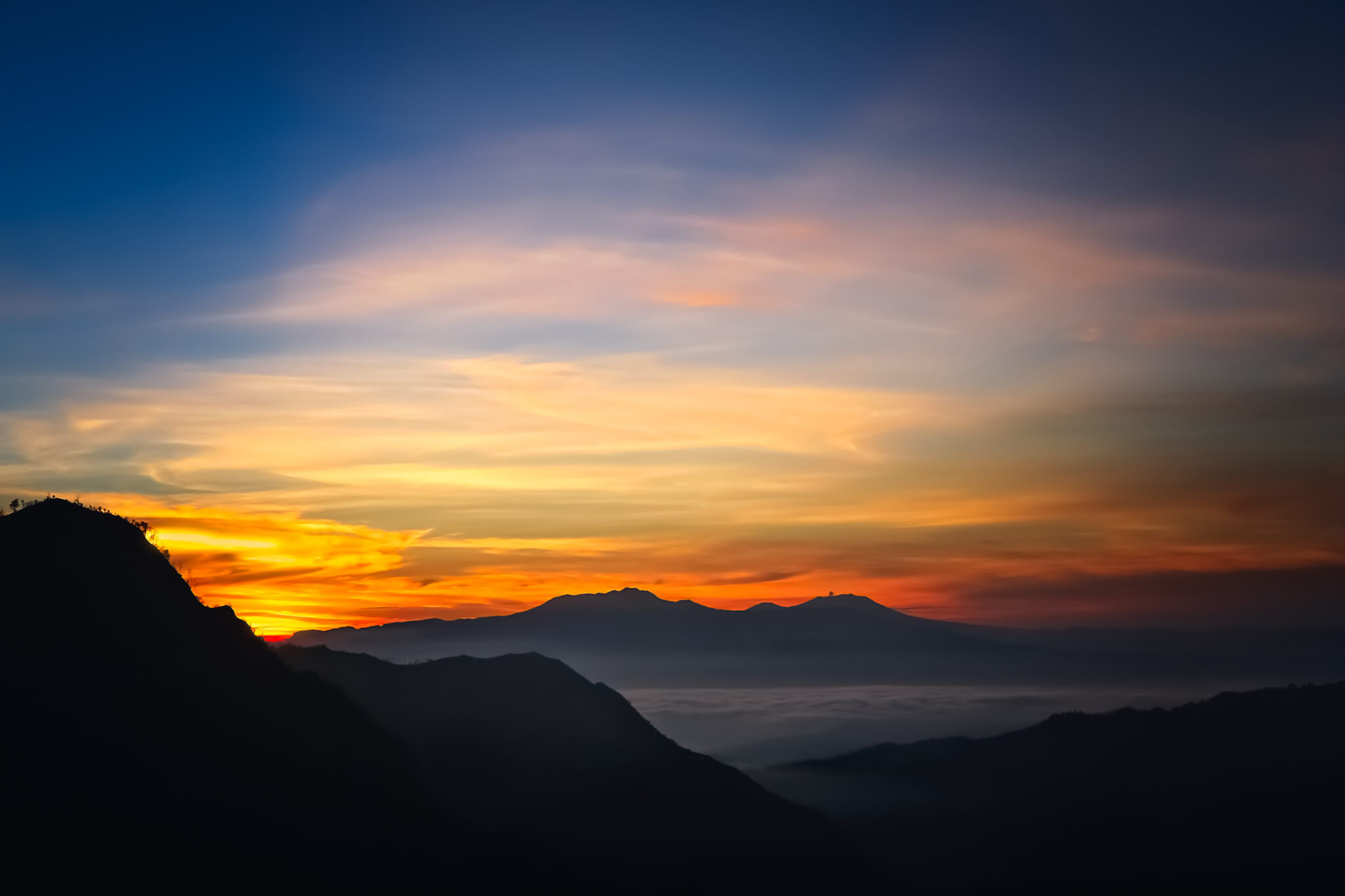 Gunung Bromo bei Sonnenaufgang: goldenes Orange am Himmel, dunkle Bergsilhouetten und Nebel über den Tälern, klare Ruhe in der Landschaft
