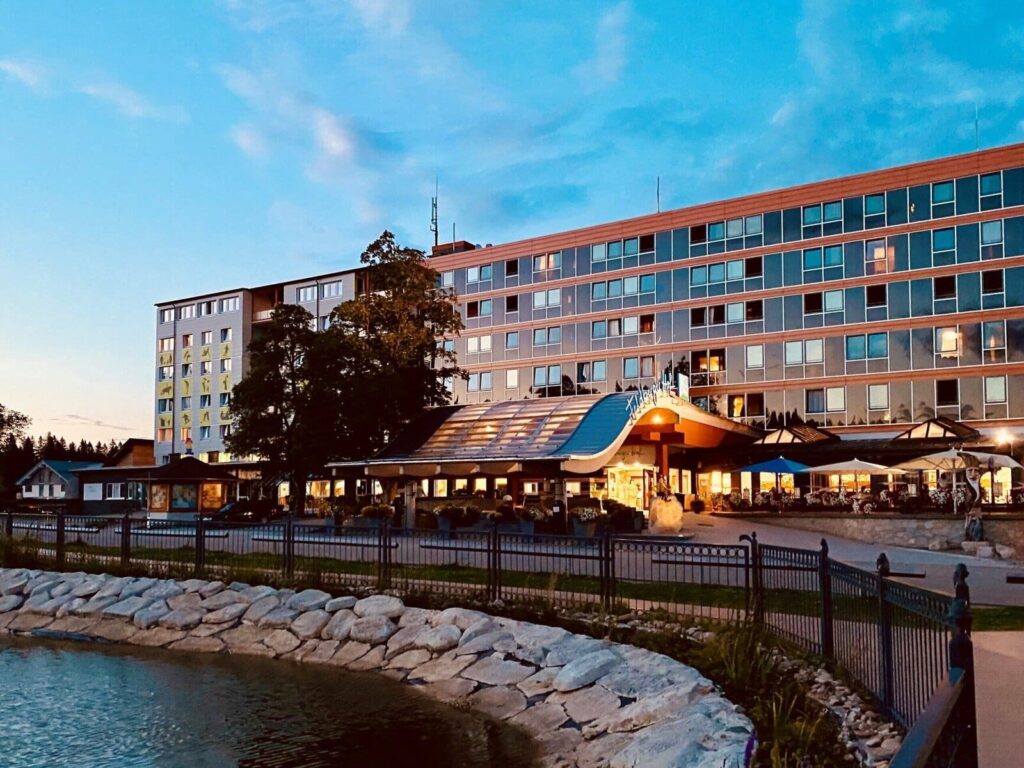 Feldberger Hof: mehrstöckiges Hotelgebäude mit rotbraunen Fassaden, blauen Glasflächen und überdachtem Eingang, davor Gartenweg am Wasser